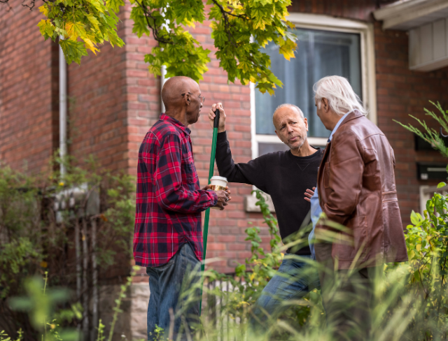 Senior Living - Aurora NWA Three older men talking outside a brick house, standing among plants and greenery. One man holds a rake and a coffee cup, while the others engage in conversation under a leafy tree.